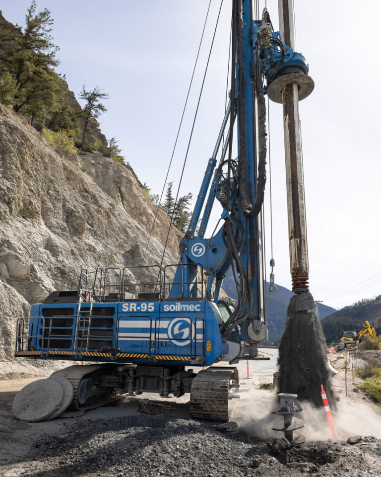 A blue drilling rig operating on a hillside, surrounded by trees and under a clear sky.