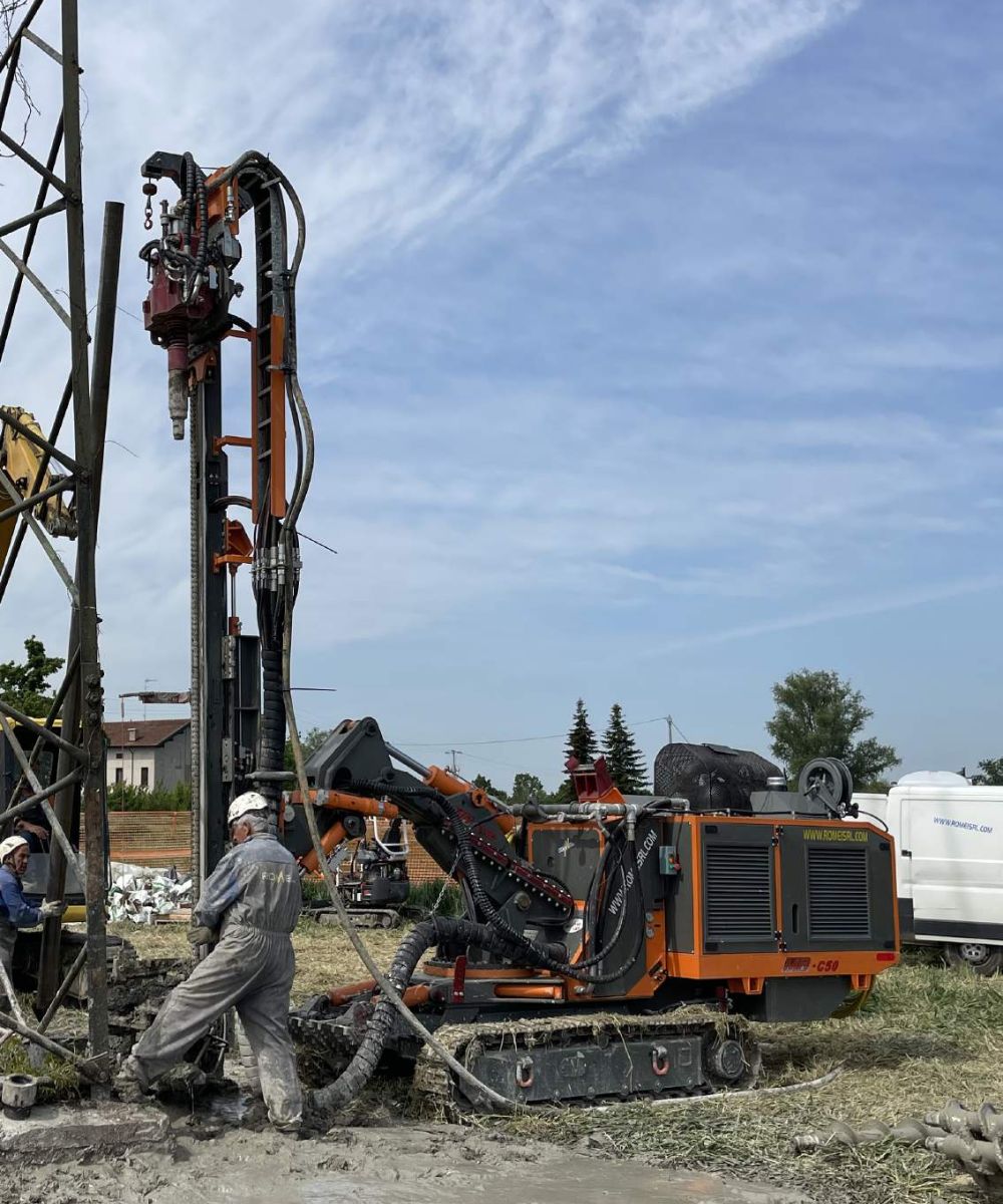 A man in orange coveralls works on a drill rig in front of a power pole.